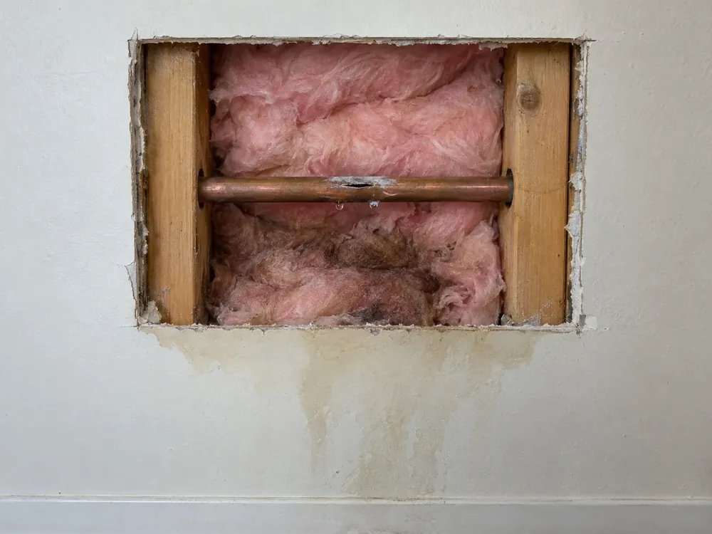 Split copper pipe with frost dripping inside an opened wall, wet insulation and water-stained drywall below